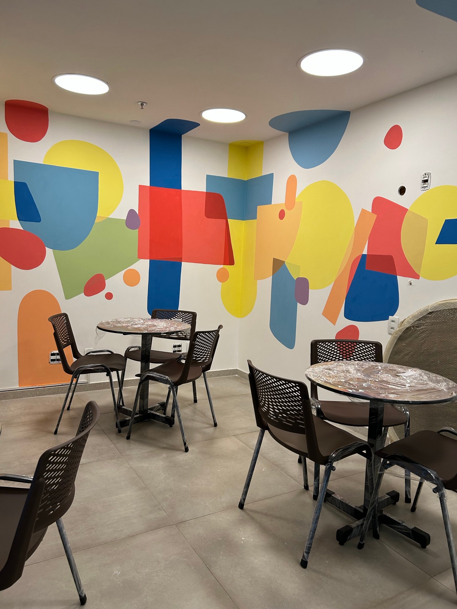 Ambiente interno com mesas, cadeiras e mural colorido